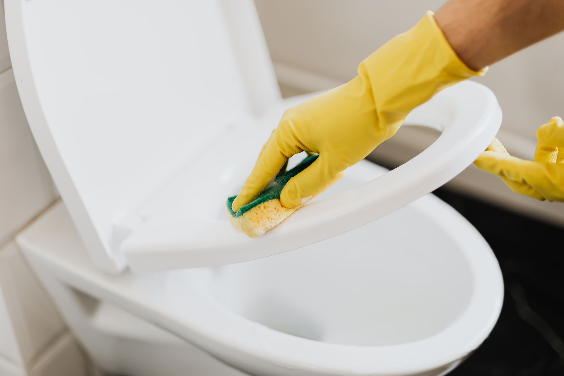From above of crop faceless person in yellow rubber cleaning gloves cleaning plastic toilet seat properly with sponge in restroom